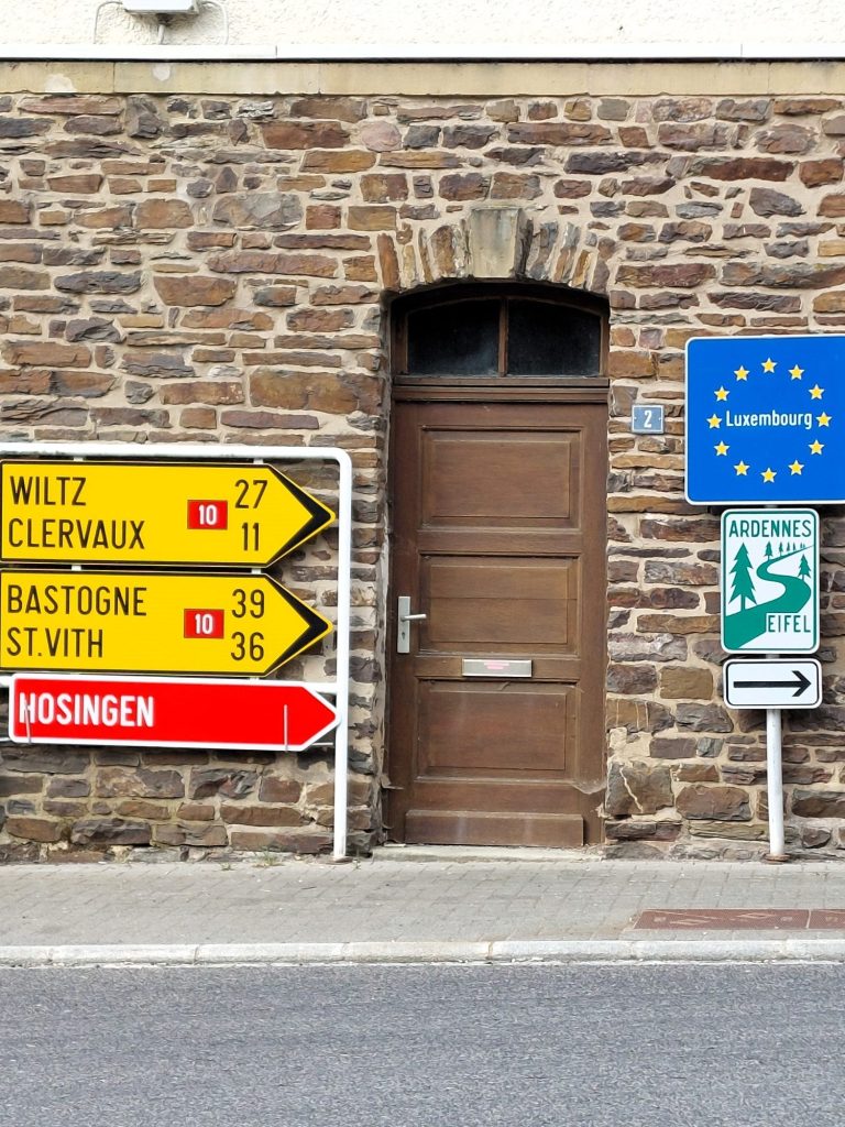 Hinweisschilder für Ortschaften und EU-Flagge Luxemburg neben einer braunen Tür an einer Steinmauer.